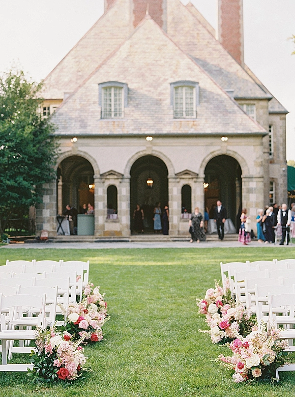 Ceremony aisle decor with white folding chairs and rose-and-greenery aisle flower arrangements on a lawn before a stone building with arches