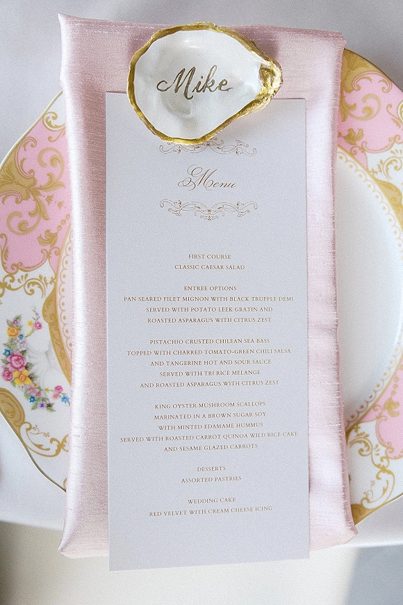 Wedding menu card with gold foil calligraphy tucked into a blush napkin on a patterned plate, paired with an oyster shell place card on a white table
