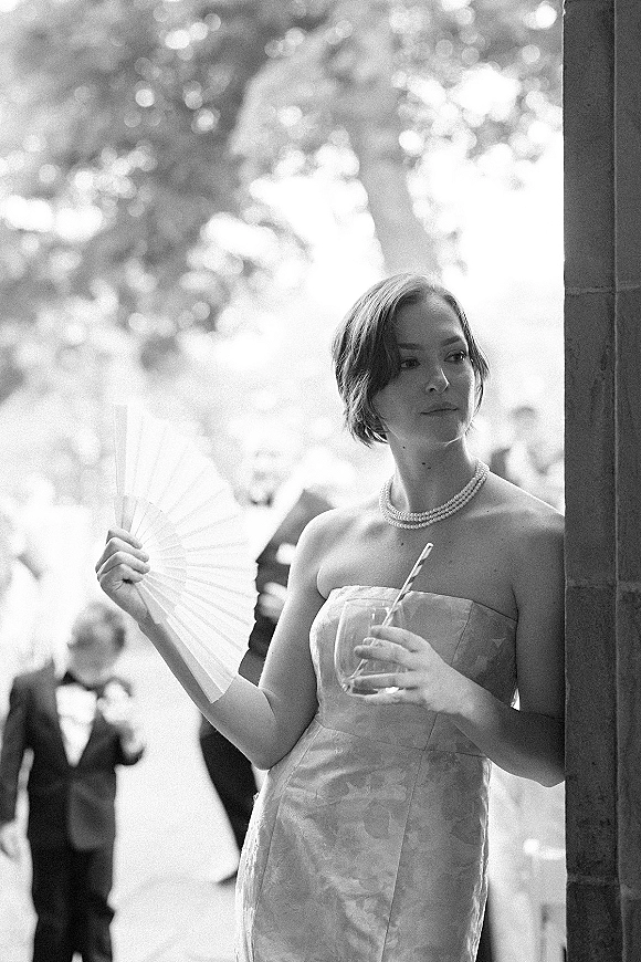Bridal portrait of a bride in a strapless gown holding a handheld fan, pearl necklace shining, with wedding guests by a stone doorway behind.