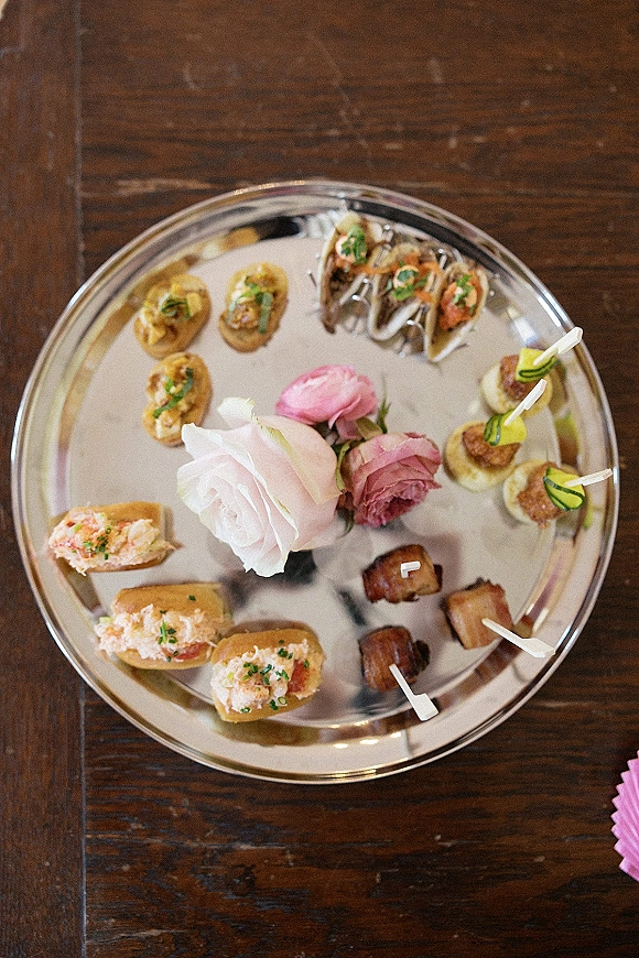 Wedding cocktail hour appetizers on a silver serving tray with crostini, mini tacos, skewers, toothpicks, and a pink rose on a wood table