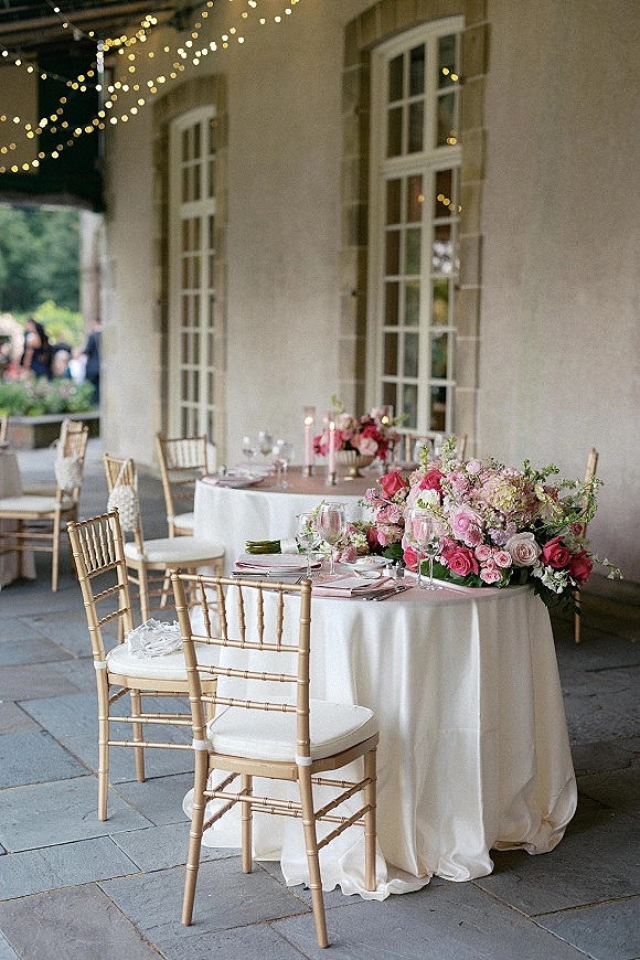 Reception tablescape with round wedding table setup, pink rose centerpieces, taper candles in glass holders, gold chiavari chairs on a terrace under string lights