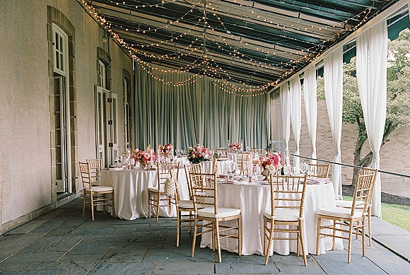 Reception tablescape with round wedding tables dressed in white linens, gold chiavari chairs, pink florals, candles, and string lights on a covered patio