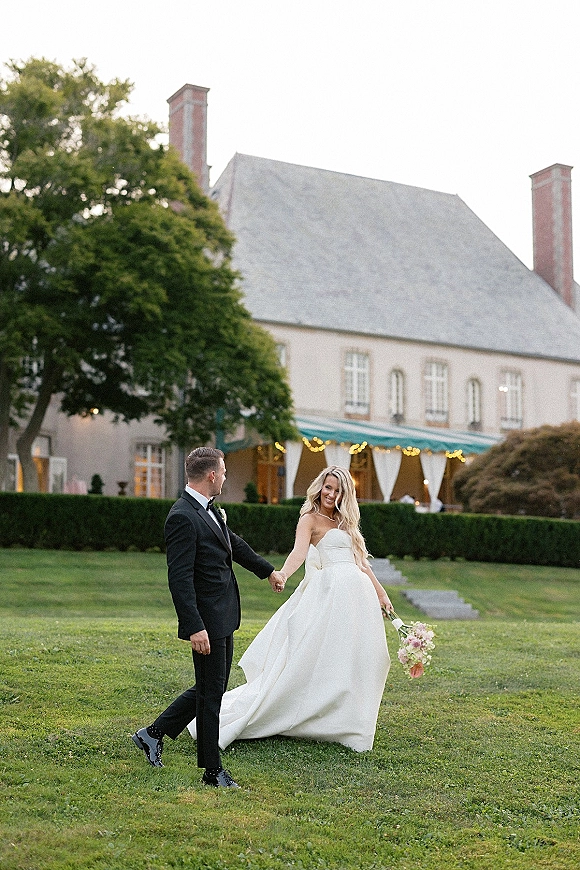 Couple portrait of bride and groom holding hands, bride with bouquet, walking on an estate lawn with hedges and a large house behind
