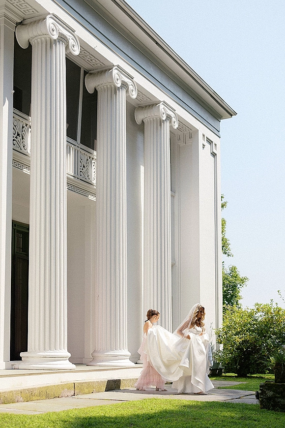 Bridal portrait of a bride with veil as a bridesmaid lifts her wedding dress train on stone steps before a white columned venue