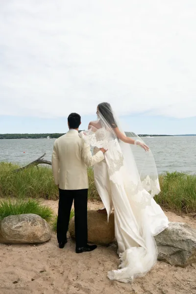 Couple portrait of bride and groom from behind, her lace veil blowing over a strapless gown as they face the ocean on a rocky beach shore