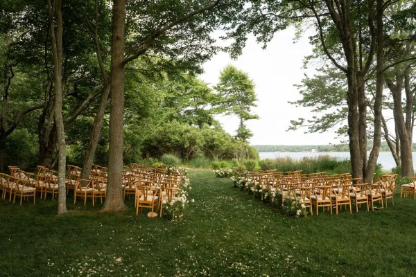 Outdoor ceremony setup with wood chairs in rows and aisle florals with scattered petals on a grass lawn by a lakeside shoreline