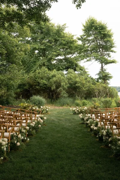 Ceremony aisle design with outdoor wedding ceremony seating, wood chairs and white florals with greenery lining a grass path on a garden lawn