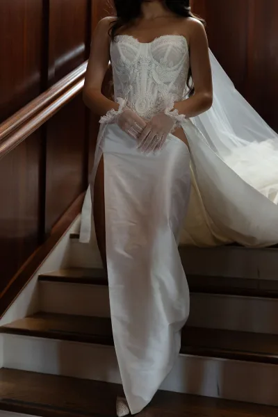 Wedding dress with a strapless corset bodice, embroidered lace, satin skirt with thigh slit, long veil and lace gloves on wooden stairs