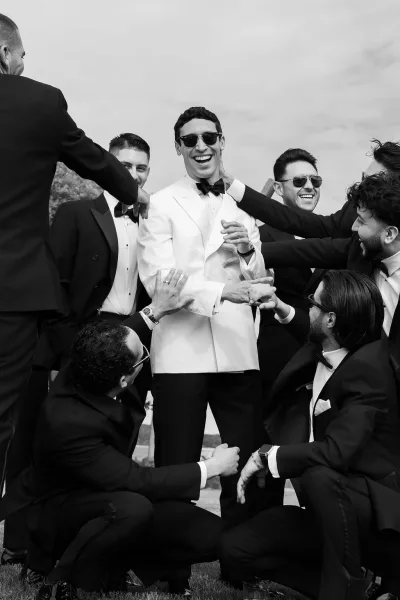 Groomsmen portrait of the groom with groomsmen in black tie tuxedos, some in sunglasses, gathered on a lawn with trees and open sky behind