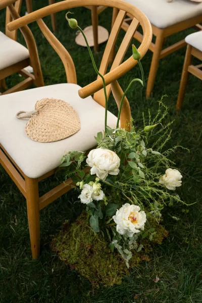 Ceremony aisle decor with wedding aisle flowers—white peonies and greenery on a moss runner beside wood chairs on a grass lawn
