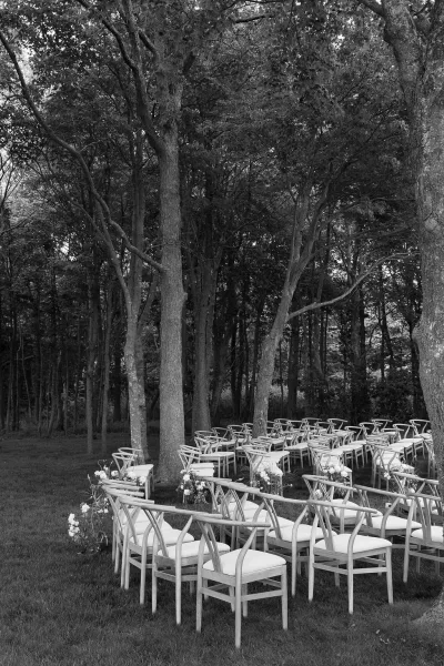 Ceremony seating with outdoor ceremony chairs, white cushions, and floral chair markers arranged in rows on a lawn in a wooded grove