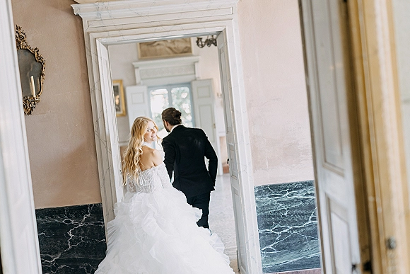 Couple portrait of newlyweds walking away as the bride looks back, long veil trailing in a bright hallway doorway with marble walls