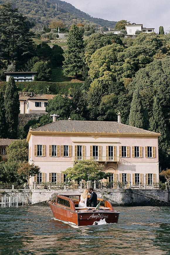 Couple on boat in a wedding boat portrait, bride in gown and groom in suit on a wooden motorboat by a lakeside villa backdrop