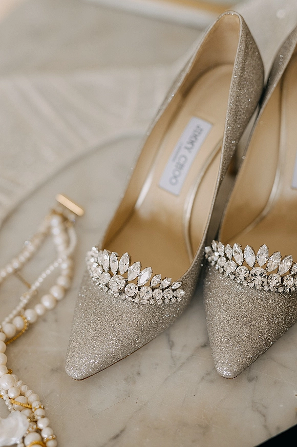 Wedding shoes with glitter heels and crystal embellishments beside pearl jewelry on a marble surface, styled in a bridal flat lay
