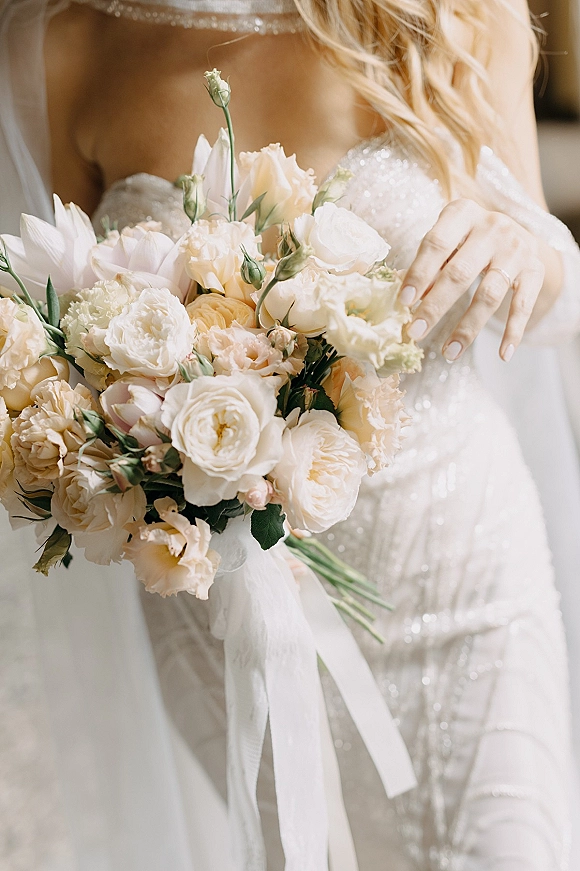 Bridal bouquet of peach and ivory wedding bouquet flowers with white ribbon and greenery, held by bride in beaded gown and veil indoors