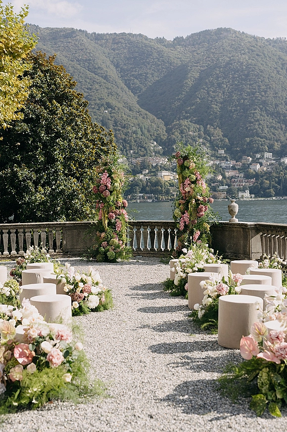 Ceremony setup with outdoor ceremony aisle lined in pastel flowers, flanked by floral pillars on a stone terrace with lake and mountains beyond