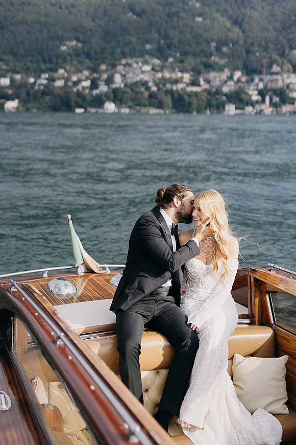 Couple portrait of bride and groom on boat, groom kissing her cheek as she smiles, mountains and lakeside town behind them