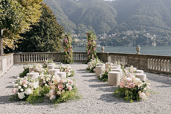 Ceremony setup with pink and white ground florals lining the aisle, framed by floral pillars and stools on a stone terrace by a lake and mountains