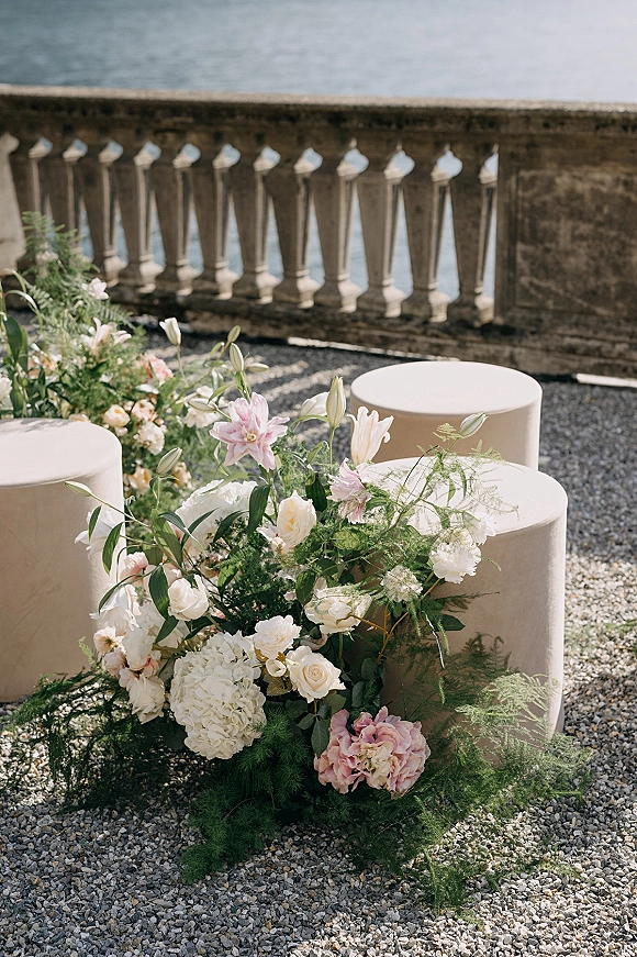 Ceremony aisle florals with a floral ground installation of white and blush lilies, roses, and hydrangea beside round beige ottomans on a gravel terrace by water