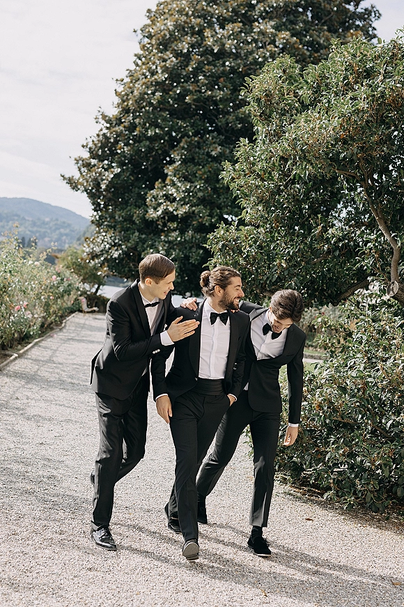 Groomsmen portrait of three men in black tuxedos and bow ties walking and laughing on a garden path with trees and distant hills
