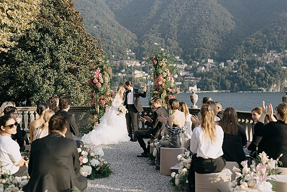Ceremony moment as bride and groom stand at a pink floral arch, bouquet in hand on a stone terrace overlooking lake and mountains
