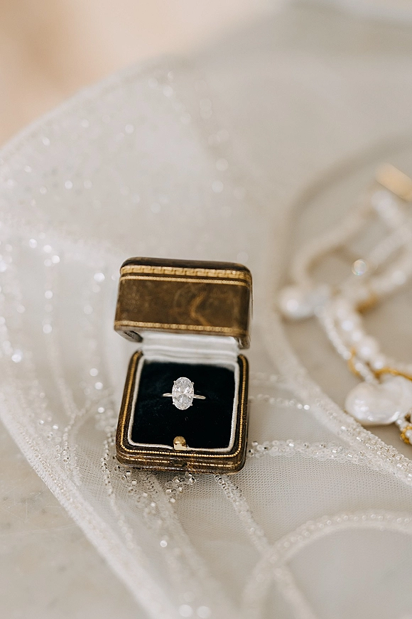 Engagement ring with an oval diamond solitaire set in a ring box, resting on sequined beaded bridal fabric on a neutral surface
