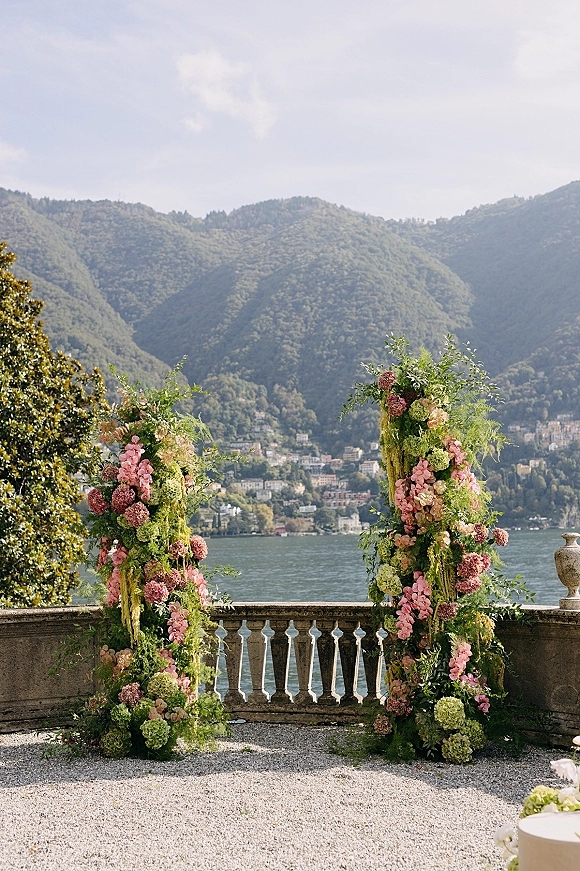 Ceremony backdrop with floral ceremony pillars of hydrangeas, pink blooms, and greenery garland on a stone terrace overlooking a lake and mountains