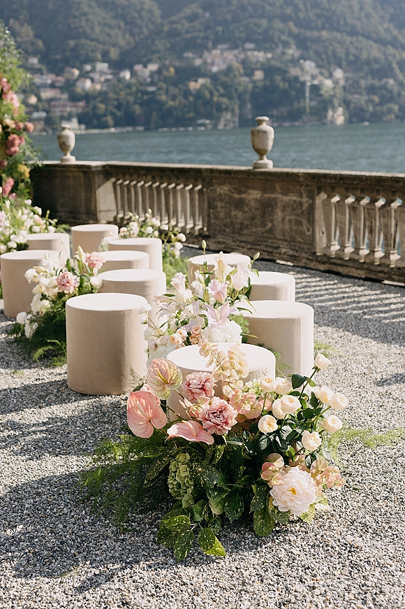 Ceremony seating with round upholstered stools beside a gravel aisle lined with wedding aisle flowers on a stone terrace overlooking lake and mountains
