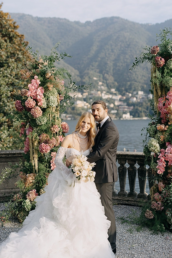 Couple portrait of bride and groom embrace beneath a pink floral arch on a stone terrace, lake and mountains behind, bride holding bouquet