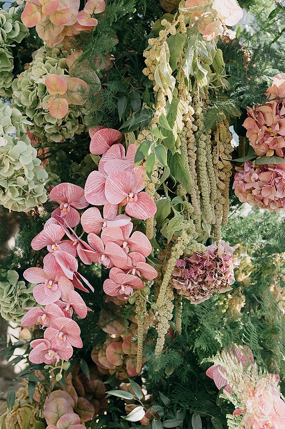 Wedding floral arch with pink orchid accents, hydrangeas, and hanging amaranthus draped over lush greenery in an outdoor garden setting