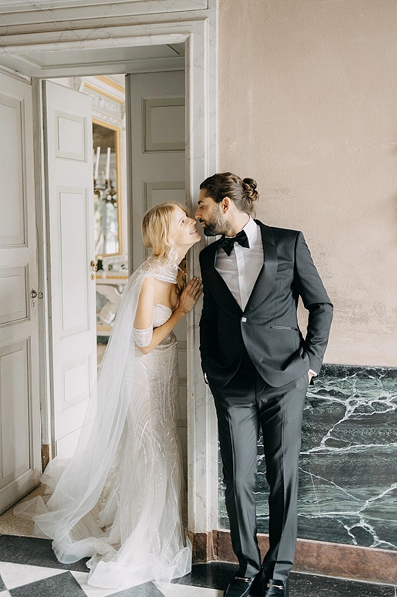 Wedding couple portrait with bride and groom doorway pose, leaning in near a kiss, her beaded gown and veil against marble wainscoting