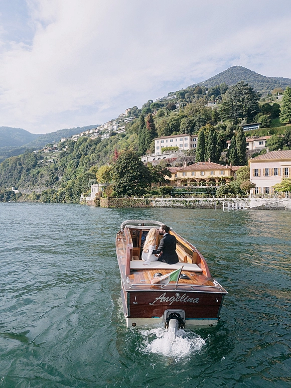 Couple on boat sharing a wedding boat ride, bride in long sleeve dress and groom in black suit on a lake with hillside villas and mountains