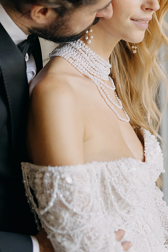 Couple portrait of groom embracing bride in lace off-the-shoulder dress, layered pearl necklace and earrings, neutral indoor backdrop