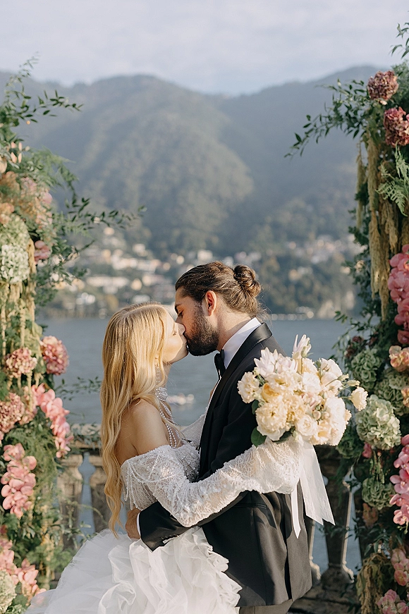 Wedding kiss as the bride and groom kissing under a floral arch, bride holding bouquet on a stone terrace with lake and mountains behind