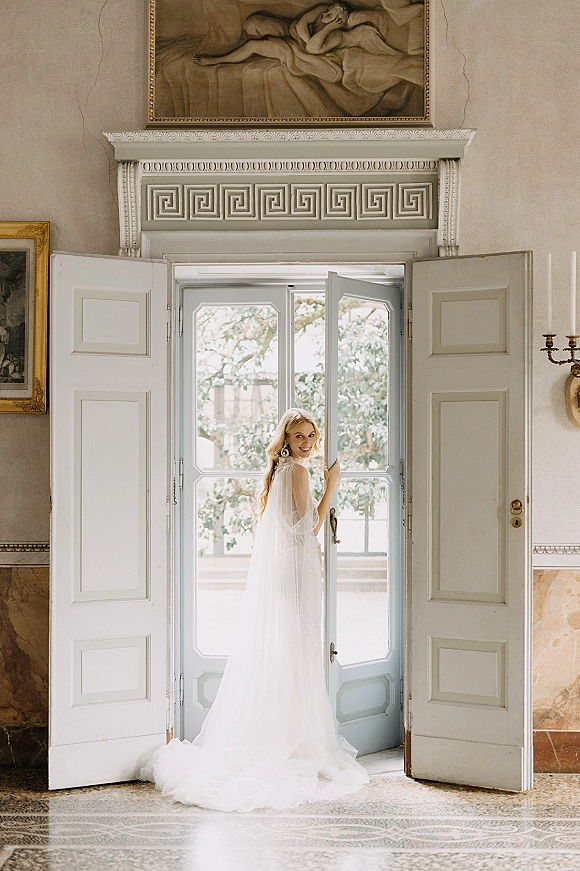 Bridal portrait of a bride at doorway looking over her shoulder, holding the door handle as window light highlights her veil in an ornate interior