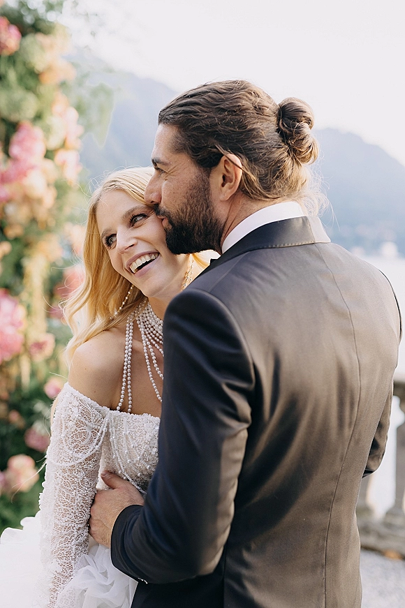 Couple portrait of bride and groom embrace as he kisses her cheek, lace sleeves and pearl necklace under a floral arch by mountains and lake