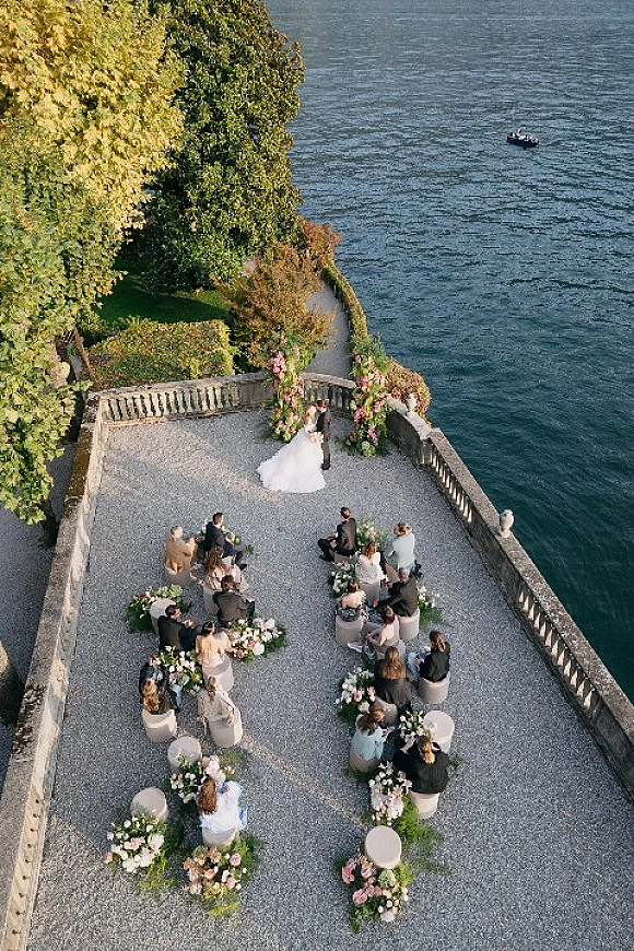 Outdoor wedding ceremony with bride and groom under a floral arch on a stone terrace by the lake, guests seated in a semicircle