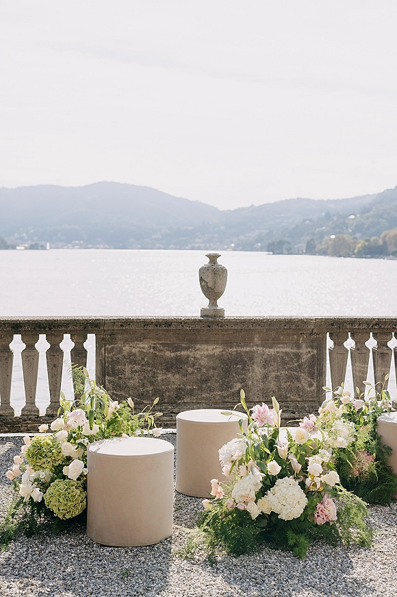 Ceremony decor with ground floral arrangements of roses, hydrangeas, and greenery around round plinths on gravel, set on a lakeside stone terrace