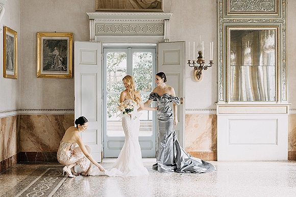 Bride getting ready as bridal party helps adjust her wedding dress train, holding bouquet in an ornate interior with mirror and doors