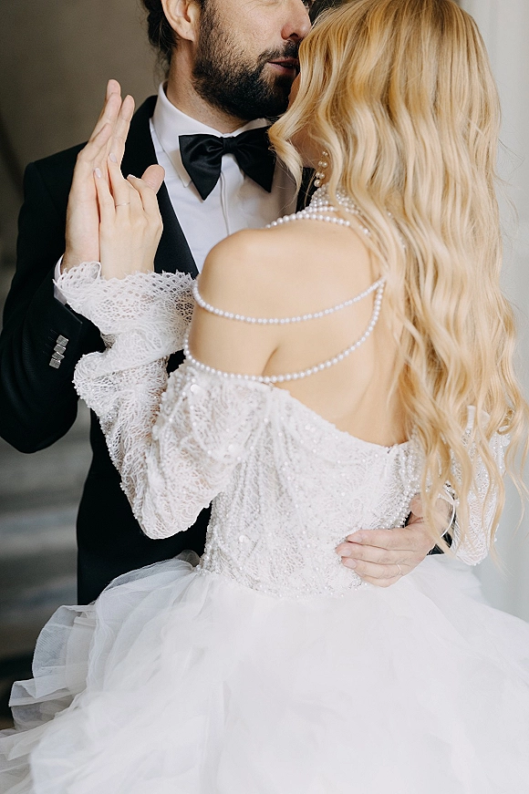 Wedding couple portrait of bride and groom embrace, her ring hand on his chest, pearl straps and black tuxedo against a neutral wall