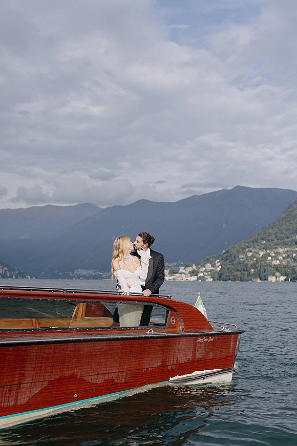 Wedding kiss portrait of bride and groom kissing on a wooden boat, bride in lace gloves and pearls on a mountain lake under clouds