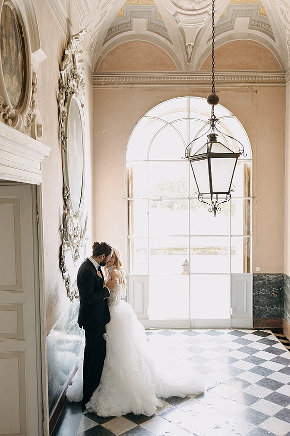 Wedding couple portrait of bride and groom kiss beside an ornate mirror in a grand hallway with arched window and checkered marble floor