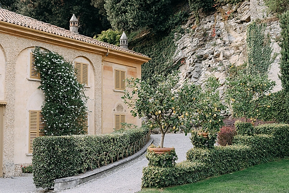 Wedding venue exterior with lemon trees lining a gravel path, ivy-covered shutters and terracotta roof tiles against a stone cliff backdrop