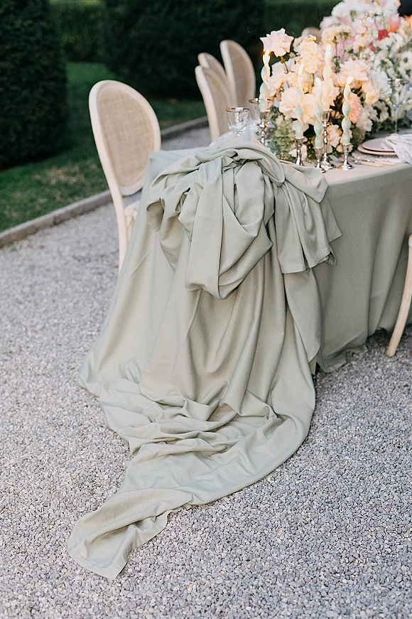 Reception tablescape with sage green tablescape linen, draped fabric bow, white floral centerpiece and taper candles on a garden gravel setting