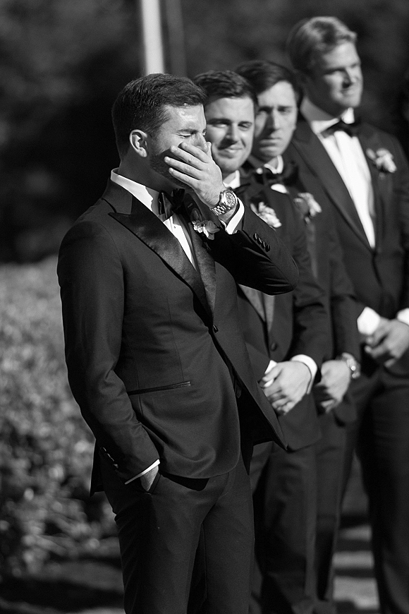 Groom reaction as he cries, covering his face in a black tuxedo with bow tie and boutonniere, outdoor greenery behind him