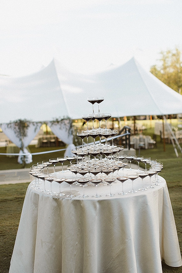 Champagne tower of coupe glasses filled with bubbly on a round white tablecloth table under a tent canopy on an outdoor lawn