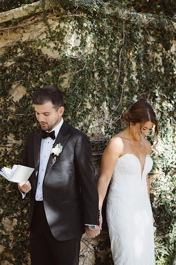 First look moment as couple holding hands while reading wedding vows from a vow book beside an ivy-covered stone wall outdoors