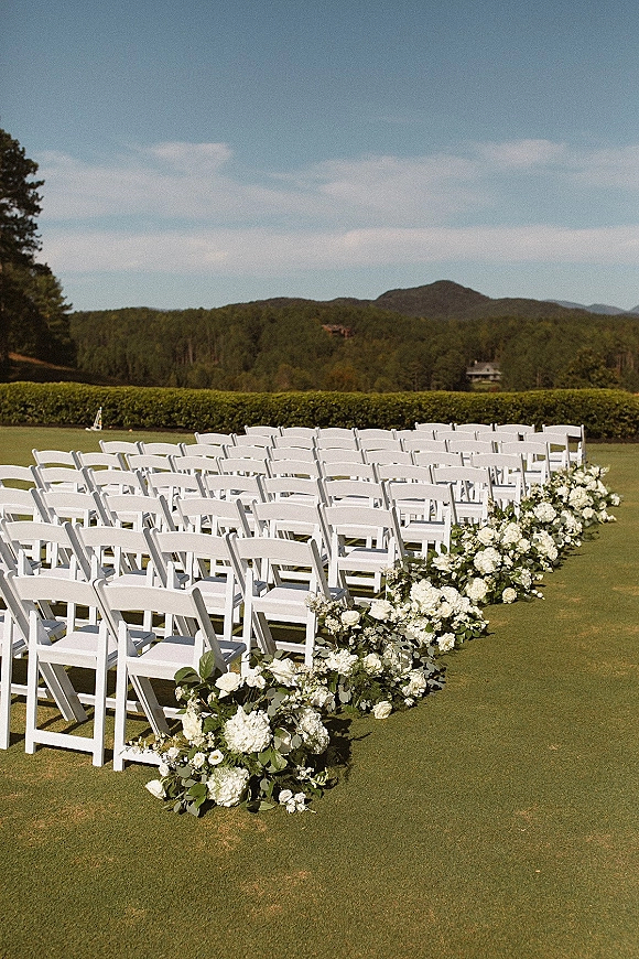 Ceremony seating setup with outdoor ceremony chairs, white folding chair rows framing a floral-lined aisle on a lawn with mountain views