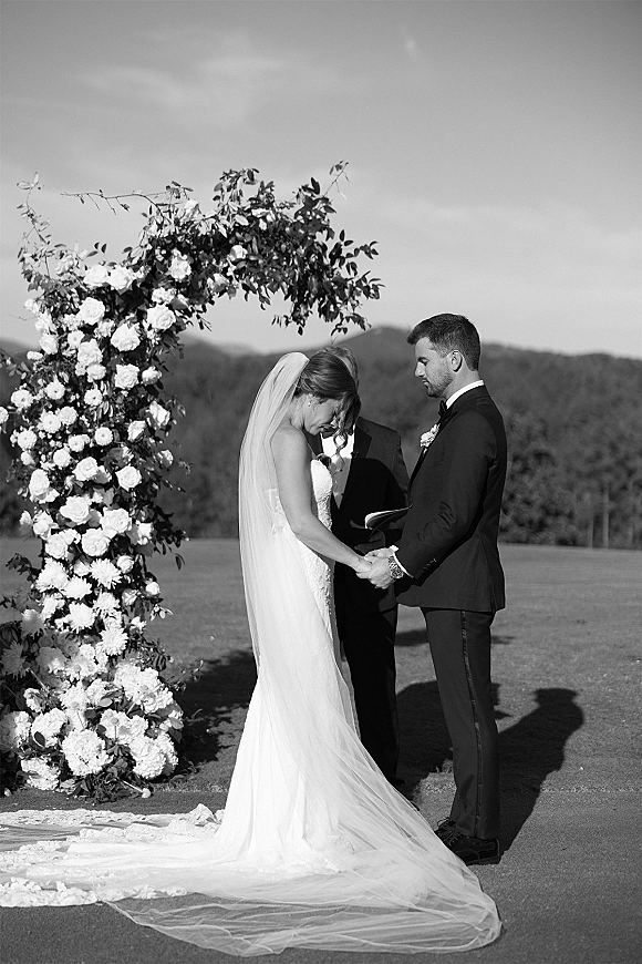 Wedding vows during an outdoor wedding ceremony as bride and groom hold hands under a floral arch with mountains behind them