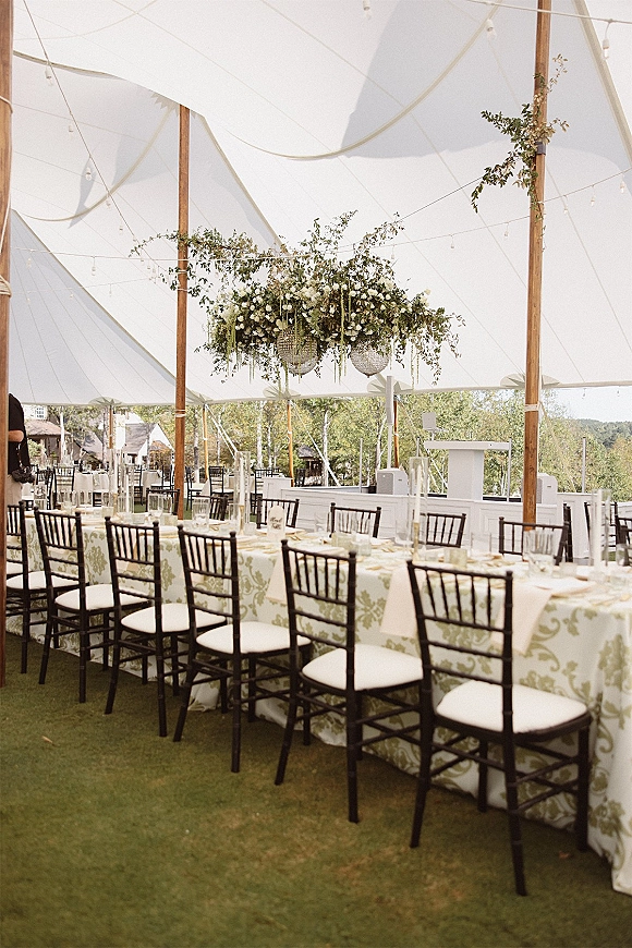 Reception tablescape with long banquet table wedding styling under a white tent canopy, with hanging greenery, string lights, and taper candles